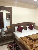 A neatly arranged bedroom with a wooden headboard, a bed with white and maroon pillows, a bedside table with a kettle, and a patterned rug on the floor.