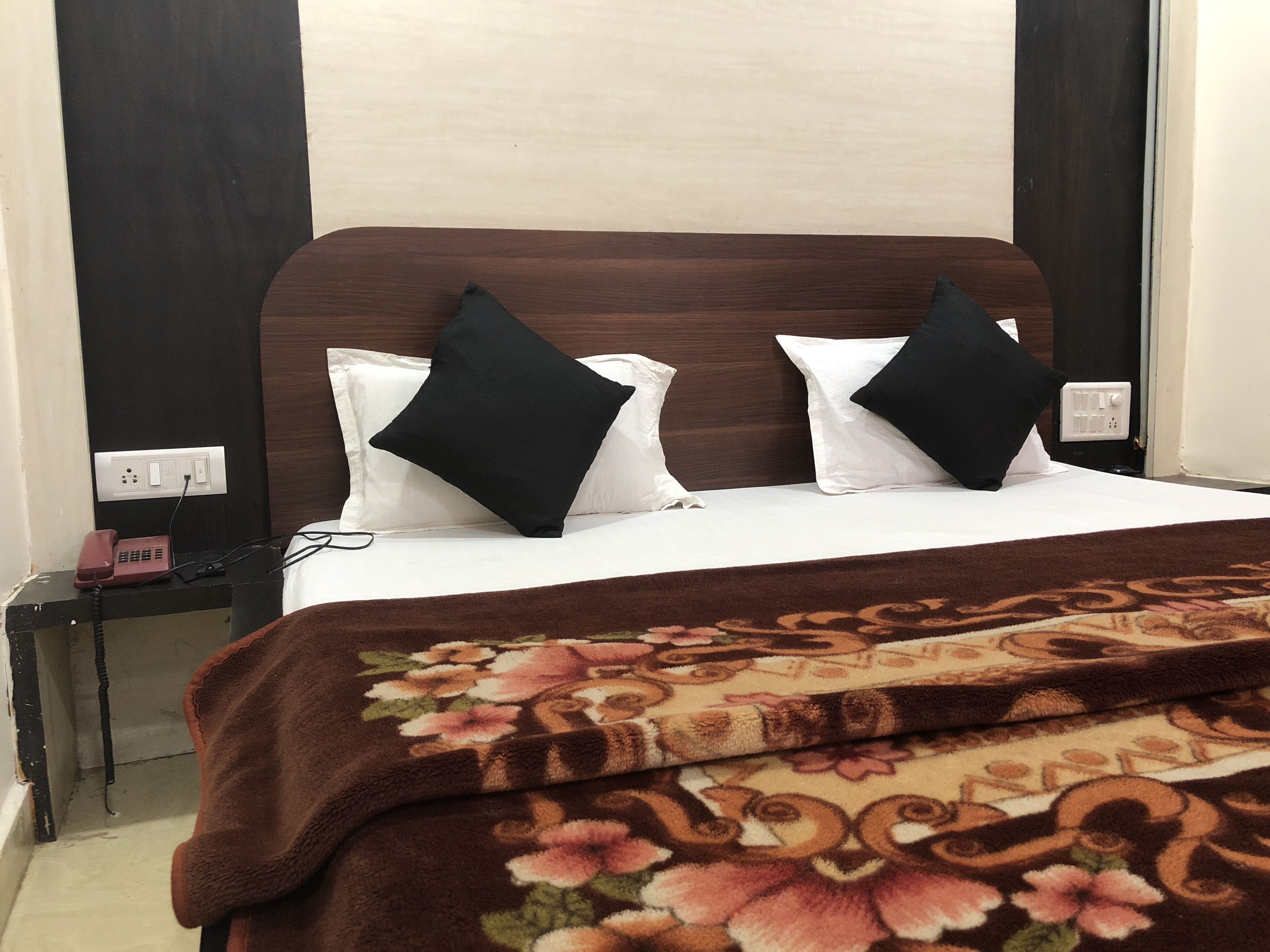 A hotel room with a bed, two black and white pillows, a brown floral blanket, a nightstand with a phone, and a wall socket.