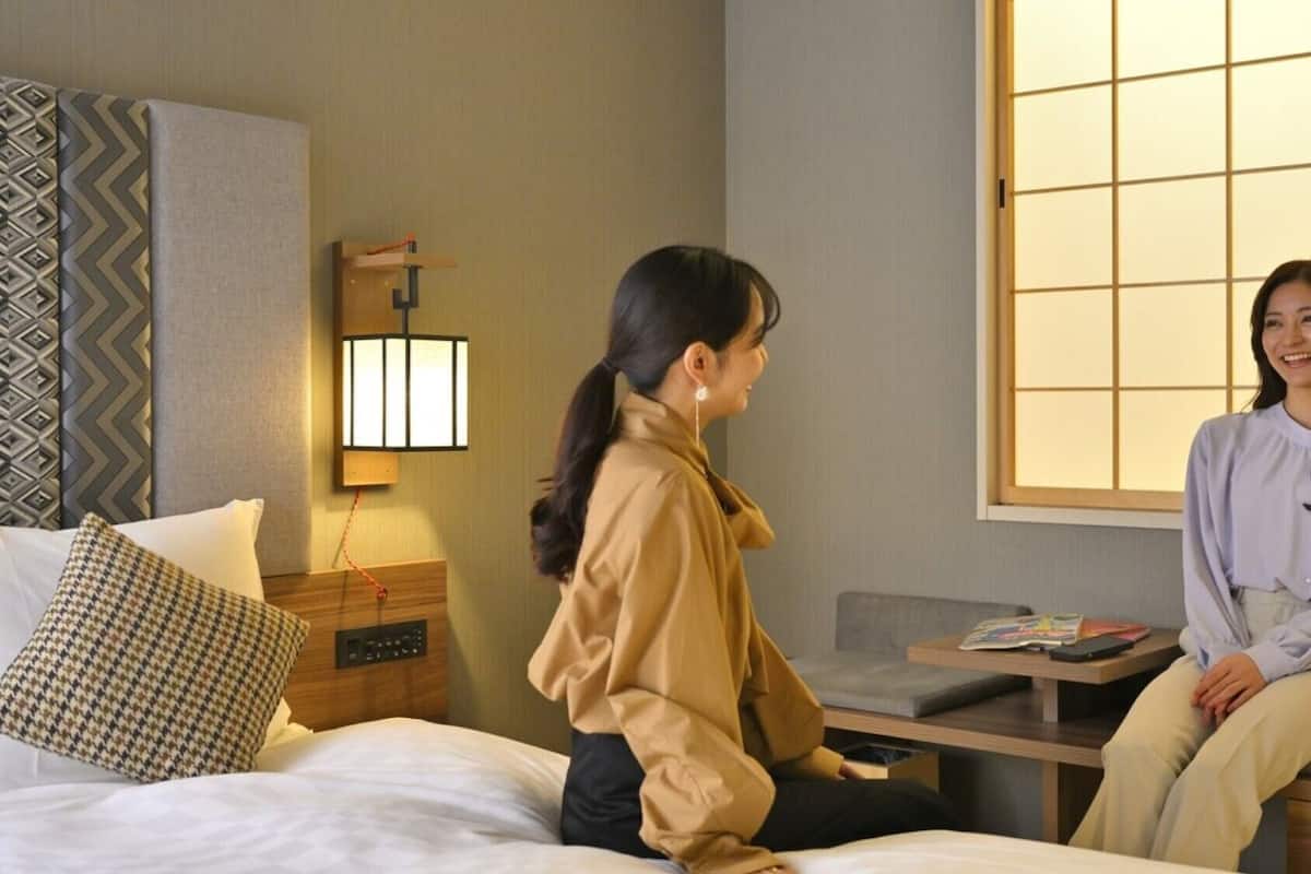 A hotel room with a bed, a bedside table, a lamp, a sofa, and a window with a view.