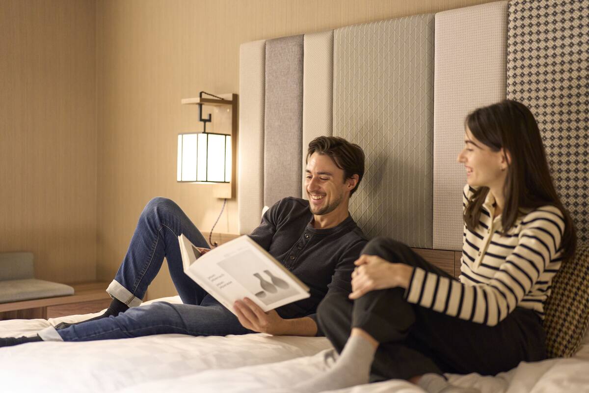 A couple sitting on a bed in a hotel room with a large window, a bedside table, and a wall-mounted lamp.
