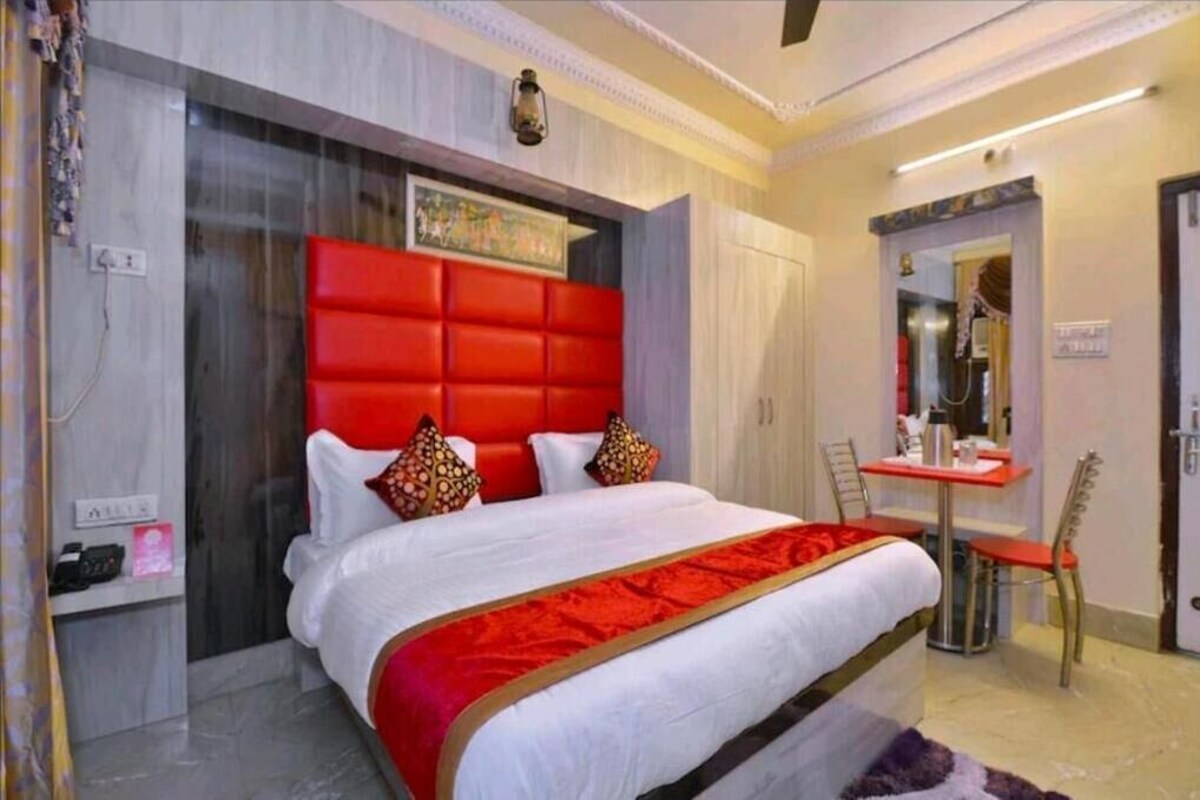 A hotel room with a large bed, a red headboard, a small desk with a chair, and a mirror.