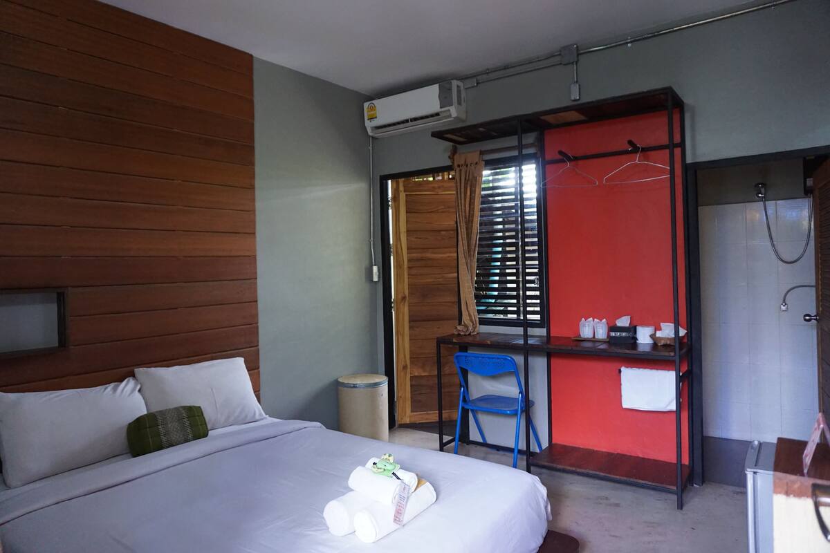 Habitación de hotel con cama, escritorio y silla, aire acondicionado, un cubo de basura pequeño y zona de ducha.