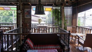 Una terraza de madera con un banco y mesas, una campana colgante, y vistas a zonas verdes y edificios.
