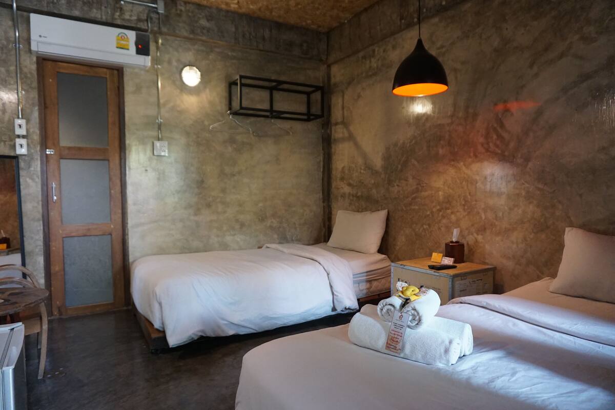 Habitación de hotel con dos camas, una puerta de madera, una mesita con una botella y una caja de pañuelos, y una lámpara colgante.