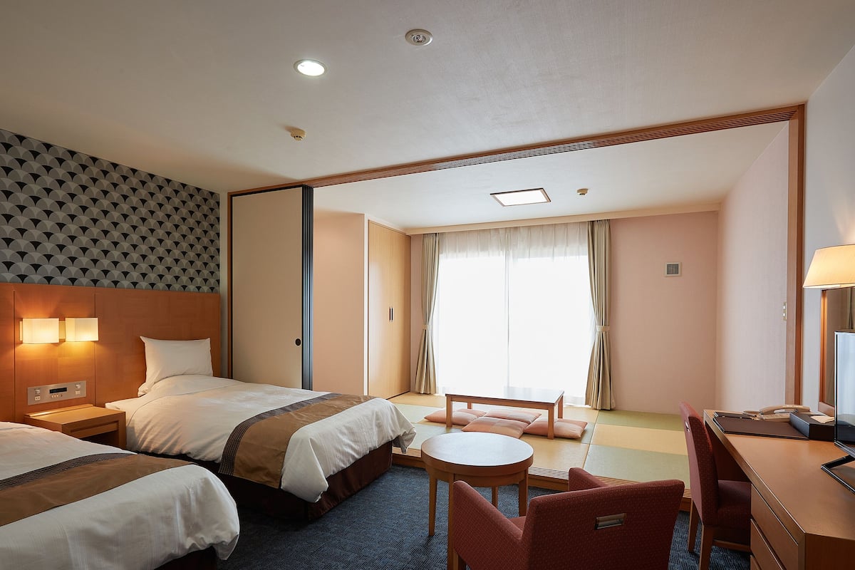Quarto de hotel com duas camas, uma escrivaninha, uma cadeira e uma mesinha.