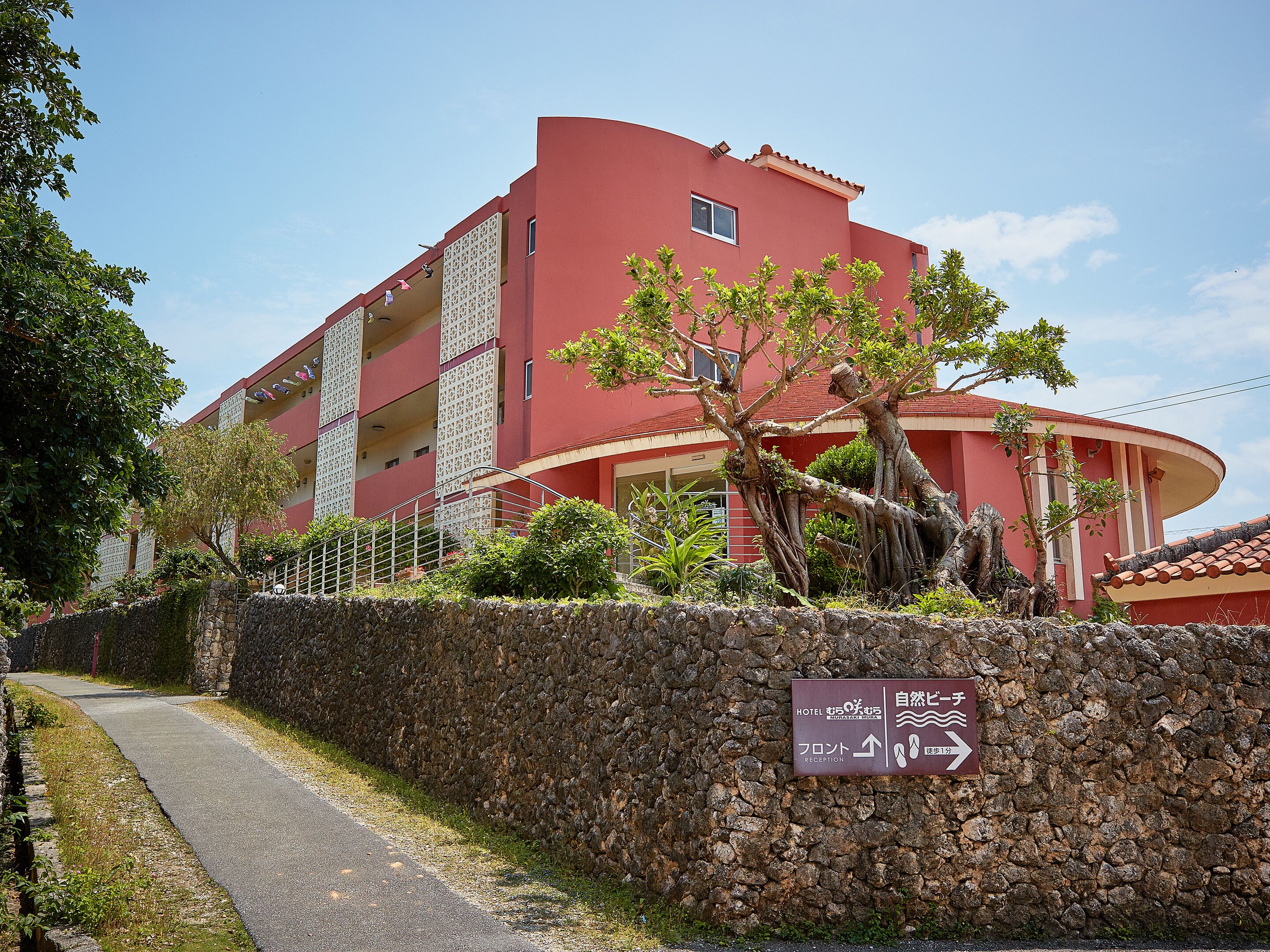 Un edificio rojo con un muro de piedra en primer plano, un letrero que indica la dirección del hotel y la recepción, y un camino que lleva a la entrada.