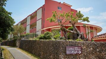 Un edificio rojo con un muro de piedra en primer plano, un letrero que indica la dirección del hotel y la recepción, y un camino que lleva a la entrada.