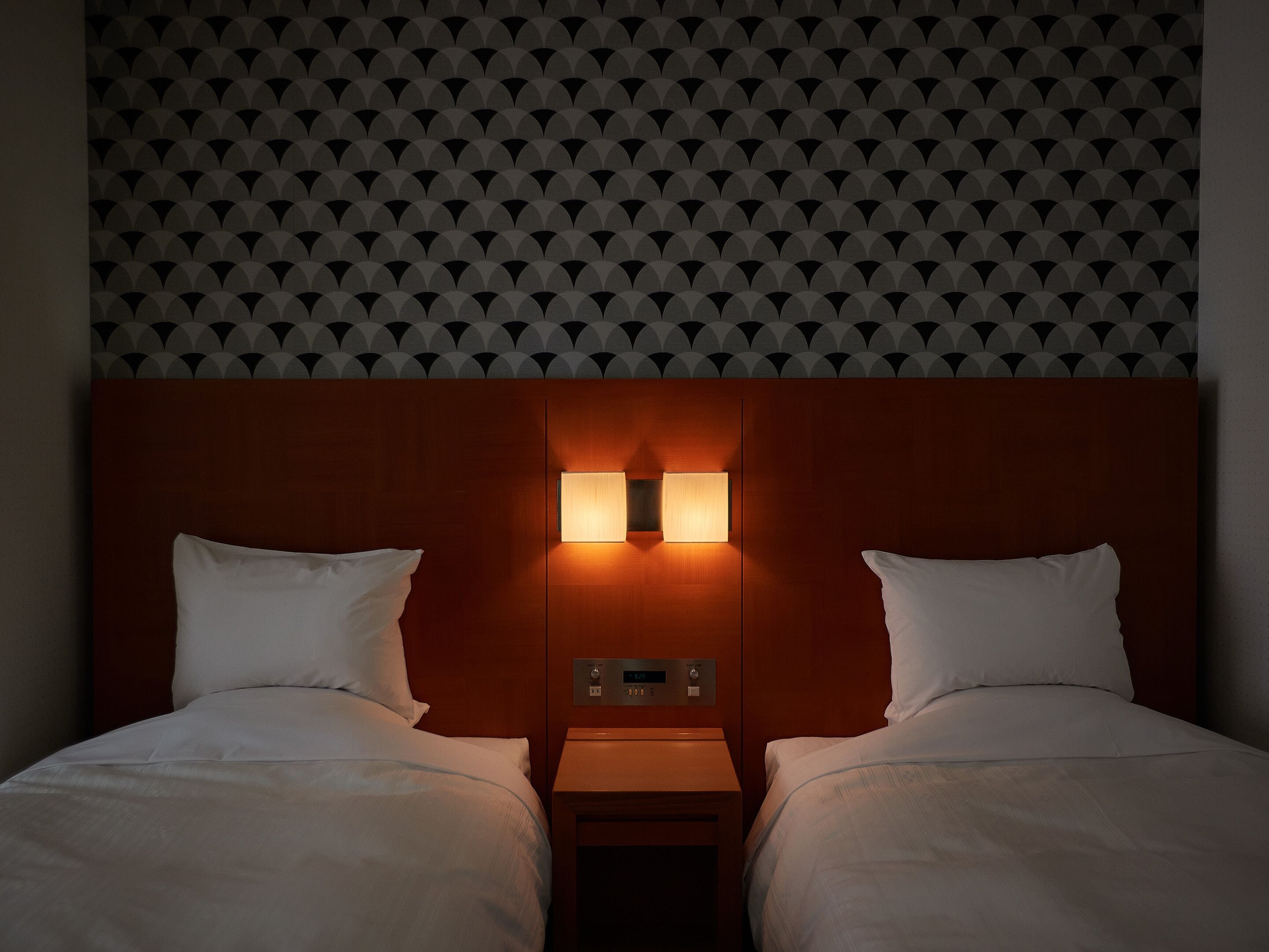 Une chambre d’hôtel avec un papier peint à motifs, une tête de lit en bois agrémentée de deux lampes murales, une table de chevet et un lit avec une literie blanche.