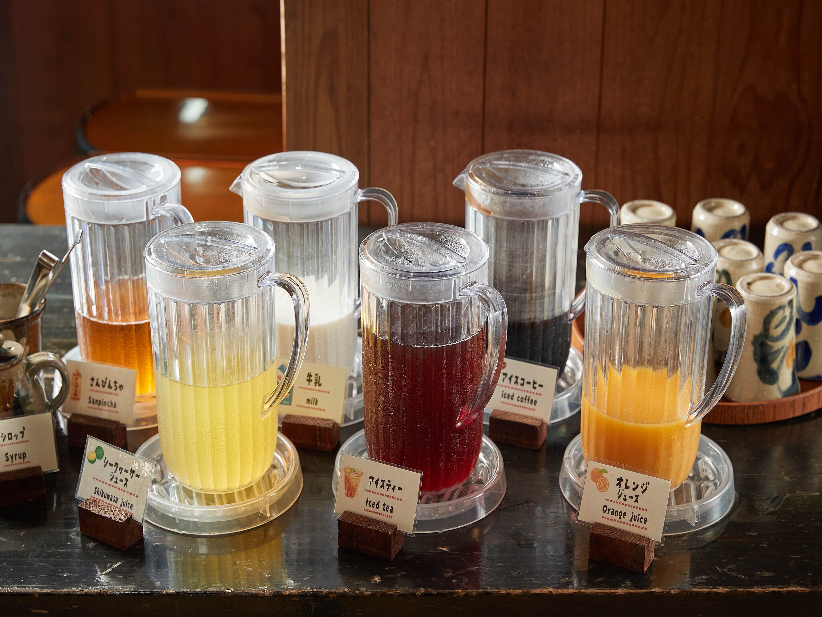 A selection of beverages in clear dispensers with labels indicating their contents, such as iced tea and orange juice, on a wooden surface.