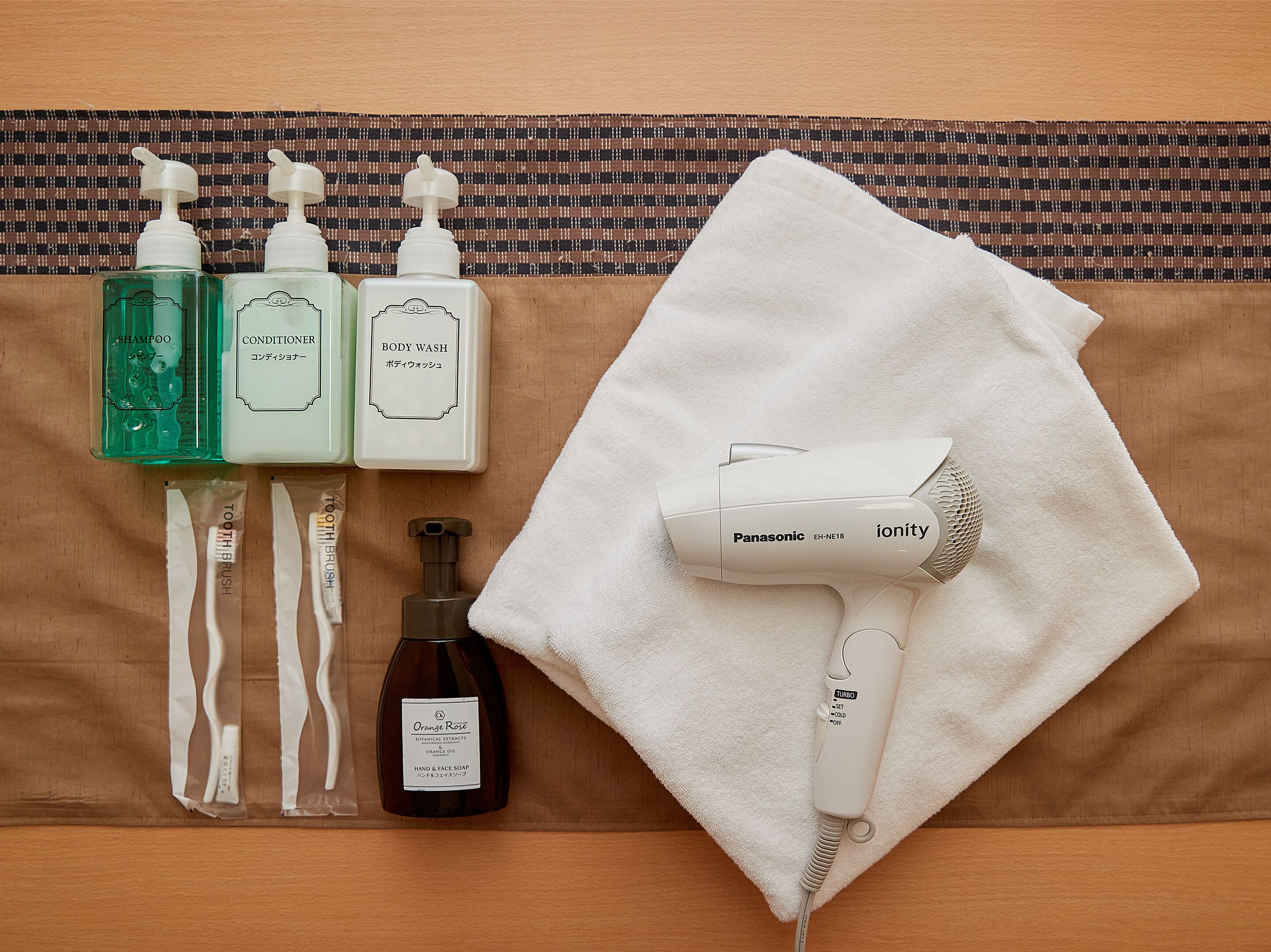 A bathroom with a towel, a hairdryer, and toiletries including shampoo, conditioner, body wash, and hand and face soap.
