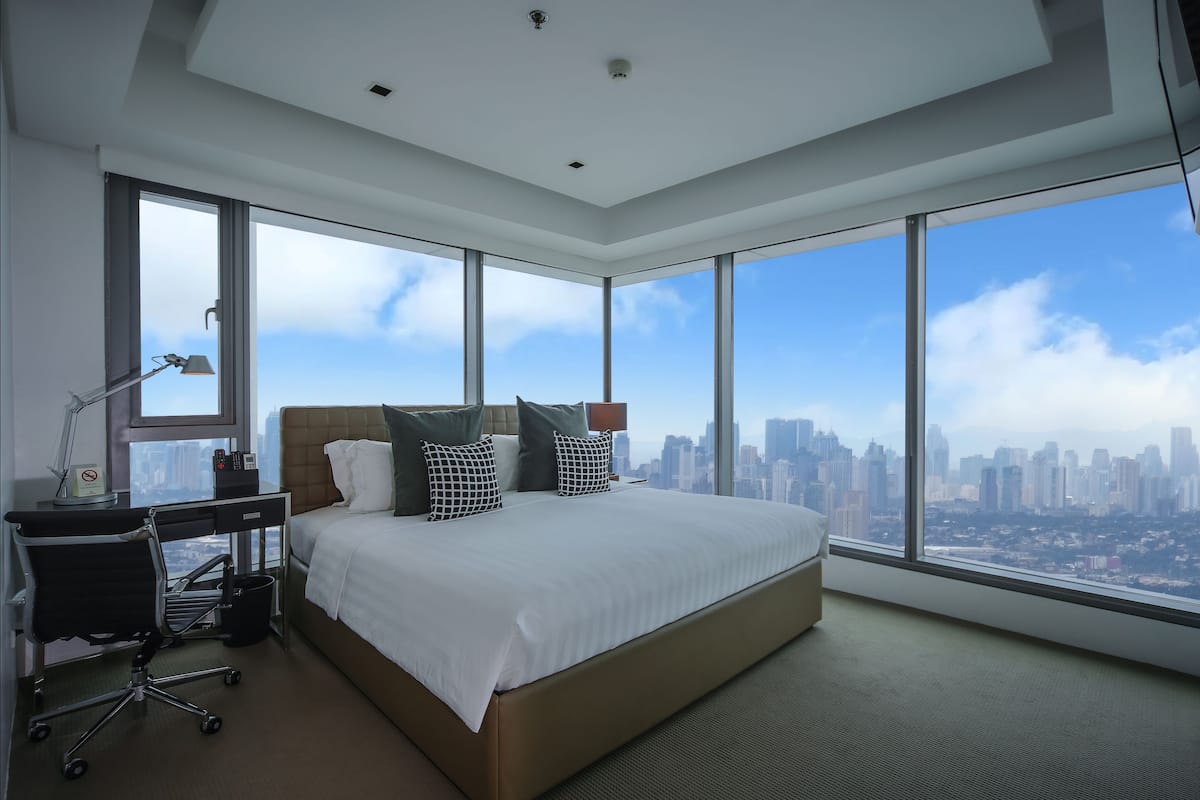 A modern hotel room with a large bed, a desk, and a chair. It features a city view through large windows.