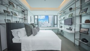 Una habitación de hotel moderna con una cama grande, estanterías empotradas con fotografías enmarcadas, un escritorio con computadora y vistas a la ciudad a través de amplias ventanas.