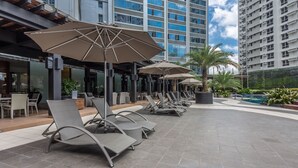 Un área de estar exterior moderna con sillones reclinables y sombrillas, una piscina y edificios altos al fondo.
