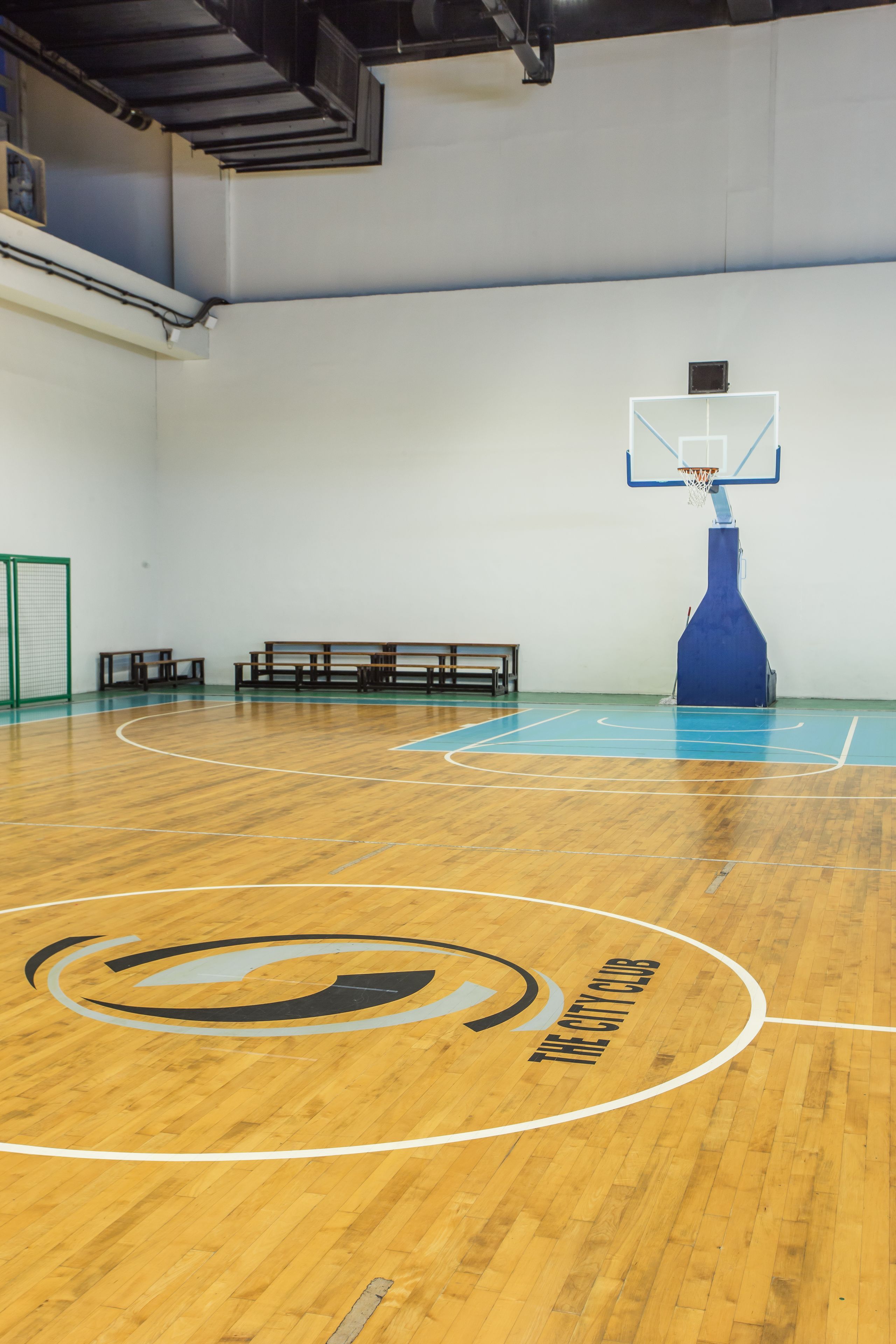 An indoor basketball court with a blue basketball hoop, wooden benches, and a logo on the floor.