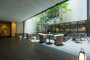 A modern indoor seating area with tables and chairs, a large window overlooking a courtyard with greenery.