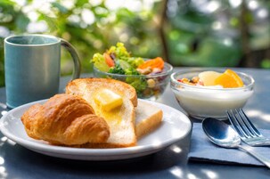 Desayuno con croissant, tostadas, mantequilla, ensalada, fruta, yogur y una taza sobre una mesa.