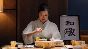 A person in traditional attire sitting at a table with bowls and chopsticks, with a framed calligraphy piece in the background.