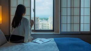 A person sitting by a window with a view of buildings, a bed with a blue blanket, and a book on the bed.