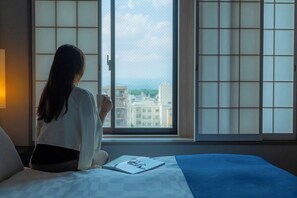 A person sitting by a window with a view of buildings, a bed with a blue blanket, and a book on the bed. - Hotel Resol Trinity Kyoto (Kyoto)
