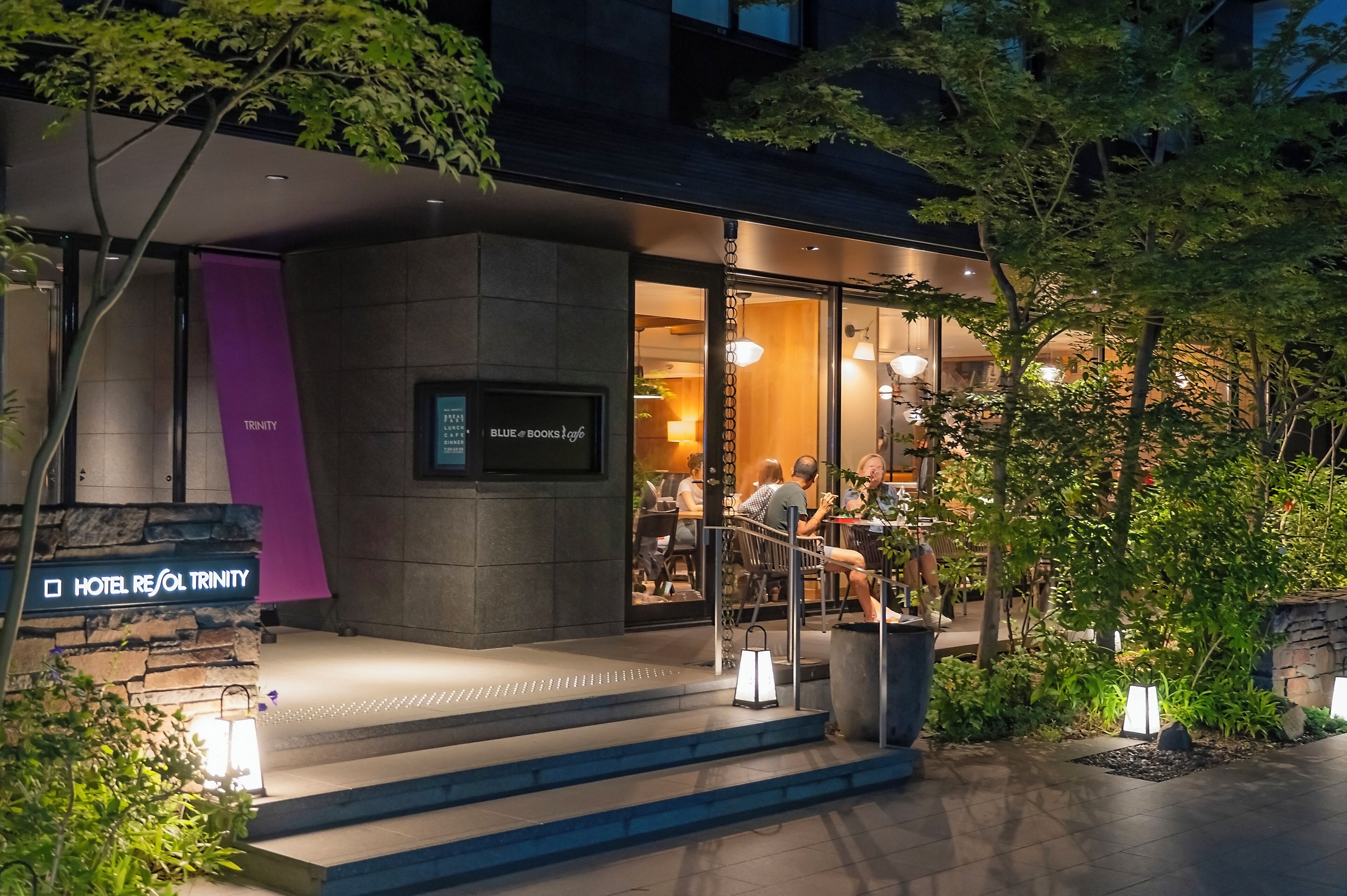 A hotel entrance with a sign reading 'Hotel Resol Trinity,' a modern exterior with glass doors, and a seating area visible inside.