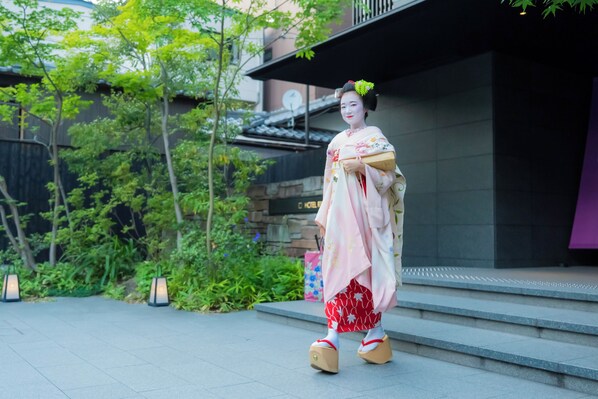 A traditional Japanese geisha in a kimono, walking on a stone pathway with greenery and a building in the background. - Hotel Resol Trinity Kyoto (Kyoto)