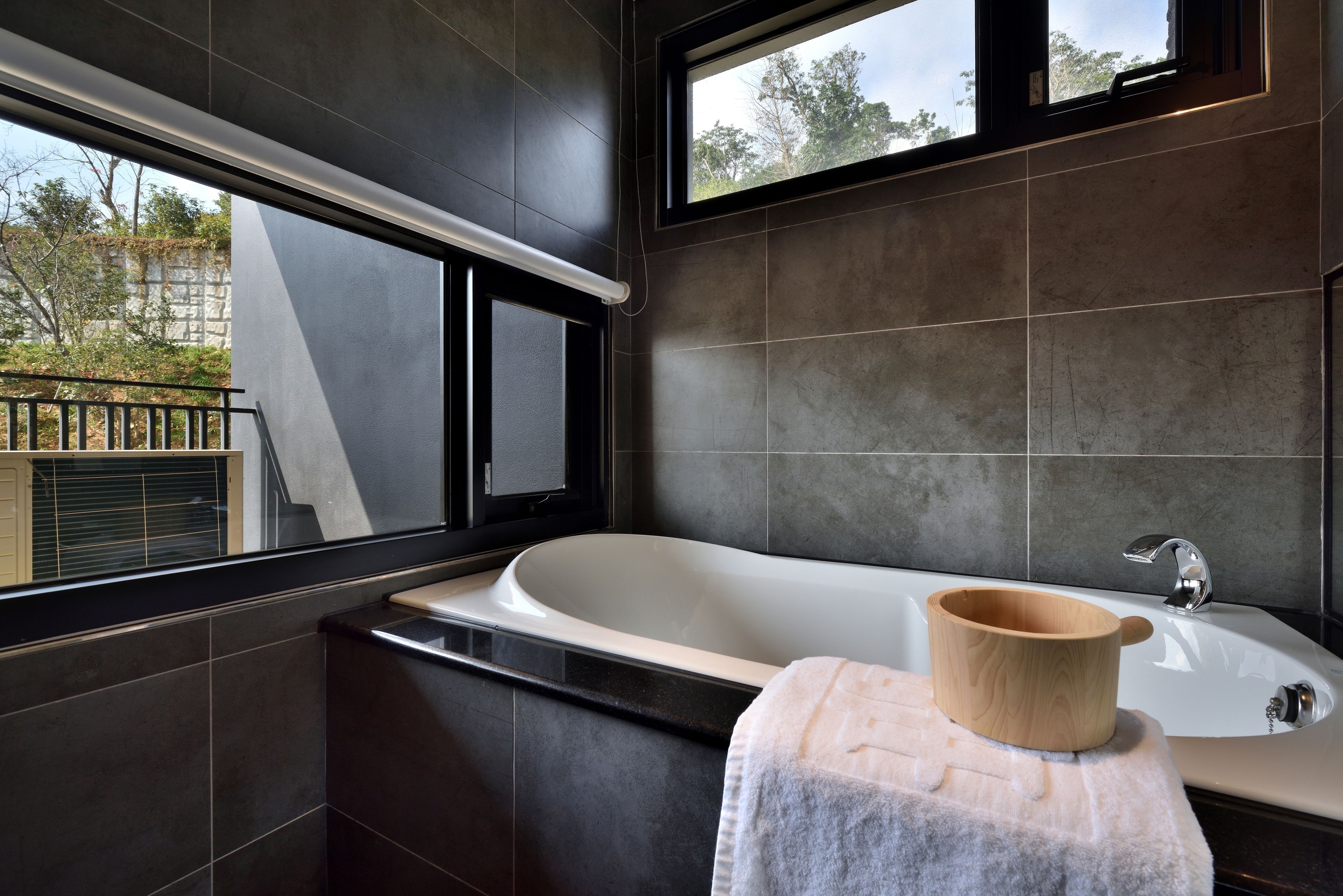 Un baño moderno con una bañera independiente, un vaso de madera sobre una toalla y una ventana con vistas al exterior.