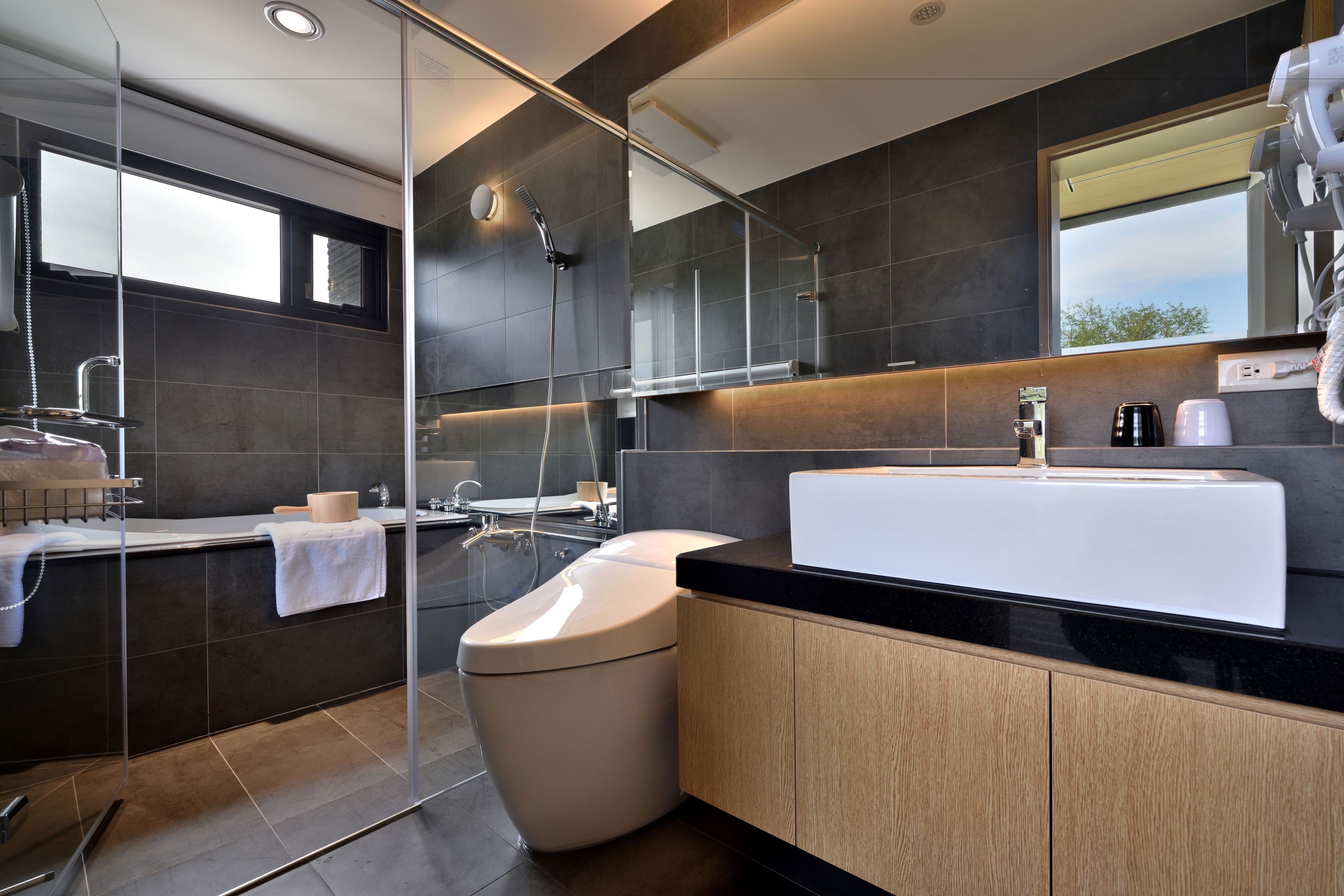 A modern bathroom with a glass shower enclosure, a white sink, and dark tiled walls.