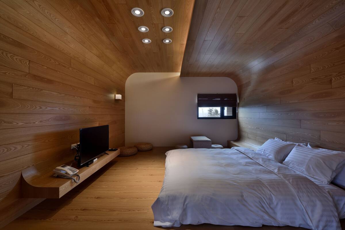 A bedroom with a wooden ceiling, a bed, a TV, and a small window.
