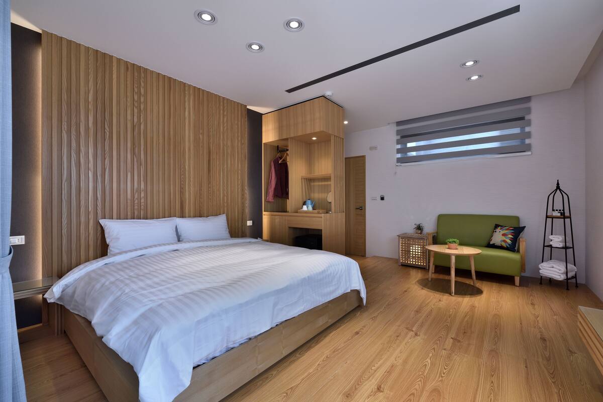A modern bedroom with a large bed, a wooden headboard, a green sofa, a small round table, and a ladder with towels.