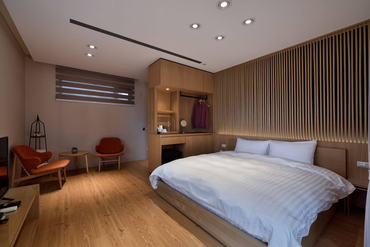 A modern bedroom with a large bed, wooden flooring, a desk with a chair, a TV, and a wardrobe.