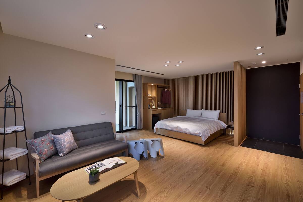 A modern bedroom with a grey sofa, a wooden bed, a small table, and a shelving unit with towels.