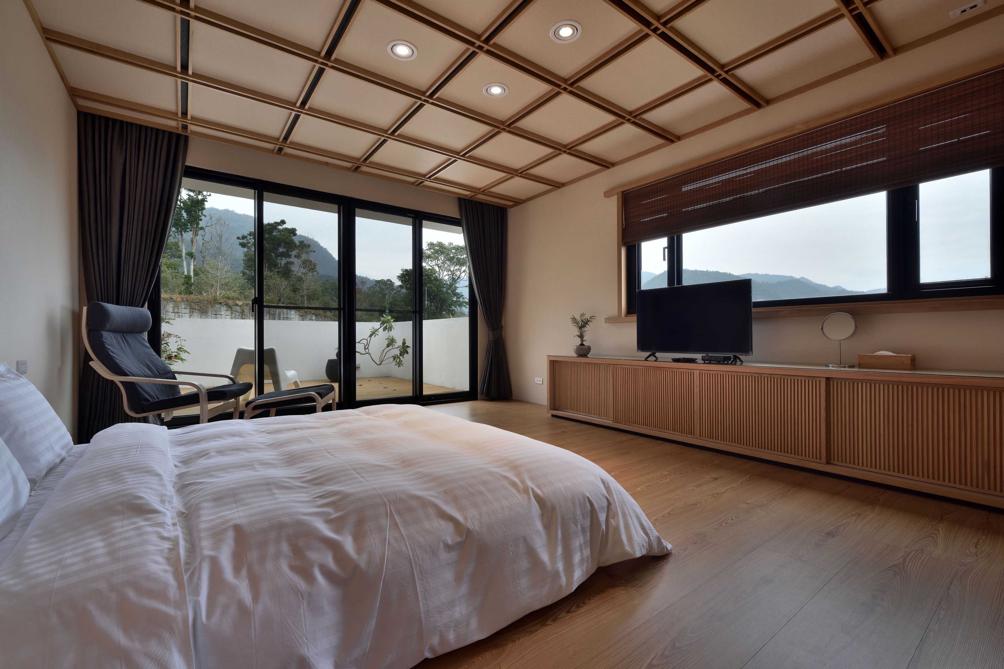 A bedroom with a large bed, a TV, a chair, and a view of the outdoors.