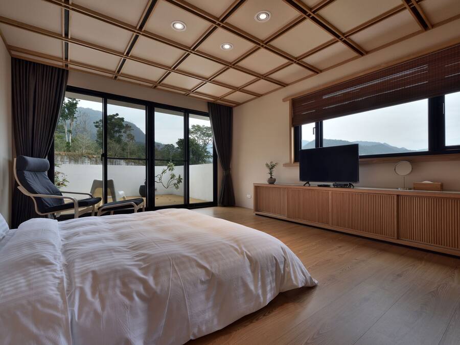 A bedroom with a large bed, a TV, a chair, and a view of the outdoors.
