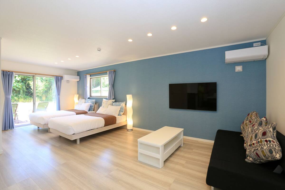 Habitación de hotel con dos camas, televisión, televisor de pantalla plana montado en la pared y un amplio ventanal con cortinas.