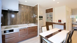 A modern kitchen with wooden cabinets, a built-in oven, and a dining area with a white table and chairs. - Stardust Villa Milky Way (Ishigaki)