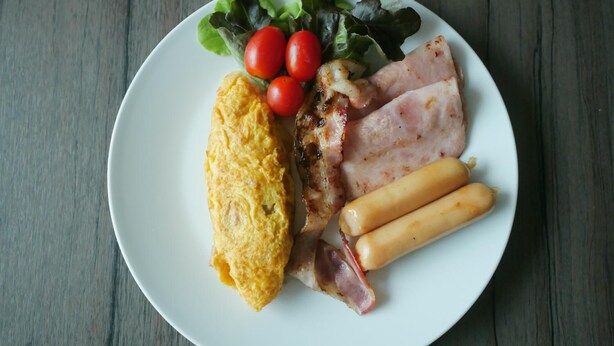 A plate with an omelette, bacon, sausages, and a side salad on a wooden table. - The Singora Hotel (Songkhla)