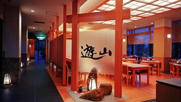 Interior de restaurante japonês tradicional, com vigas de madeira, esteiras de tatame e parede central com caligrafia.