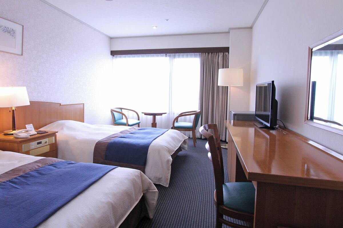 Ein Hotelzimmer mit zwei Betten, einem Schreibtisch mit Fernseher, einem Sessel, einer Lampe und einem Fenster mit Vorhängen.