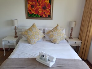 A hotel room with a large bed, two nightstands, two lamps, a painting of flowers, and a towel on the bed. - Valley Heights Guest House B&B (Cape Town)