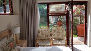 Ein Schlafzimmer mit einem Bett, einem Nachttisch, einer Lampe und zwei Stühlen, die zu einer Glastür mit Blick auf einen Garten hin ausgerichtet sind.