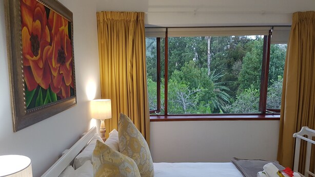 Ein Hotelzimmer mit Bett, Kissen, einer Lampe, einem Gemälde und einem Fenster mit Blick auf Grünflächen.