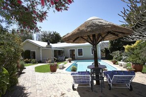 A swimming pool with lounge chairs and a thatched umbrella, surrounded by a garden and a house.