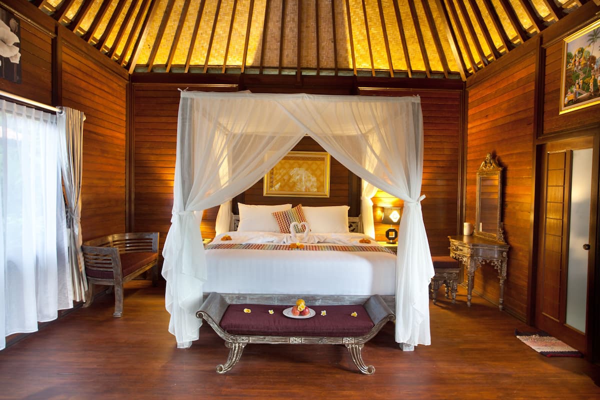 A four-poster bed with a canopy, a wooden bench with a decorative item, a wooden chair, and a nightstand with a lamp.