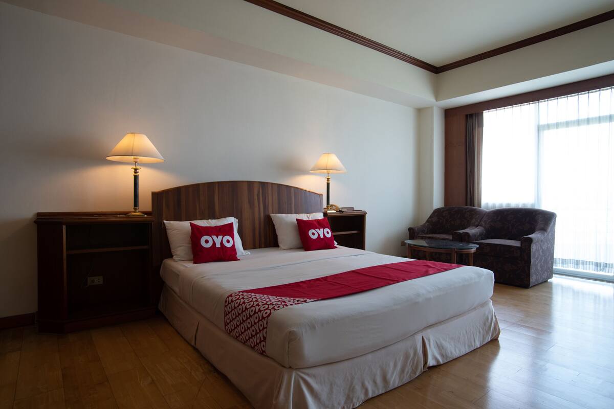 A hotel room with a large bed, two bedside lamps, a red throw blanket, a wooden headboard, a nightstand, a chair, and a large window with curtains.