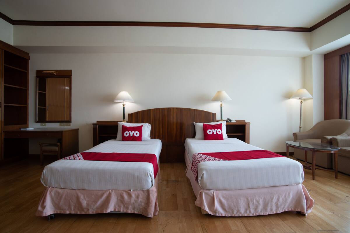 A hotel room with two beds, each with a red 'OYO' pillow, wooden headboard, bedside lamps, a desk, and a chair.