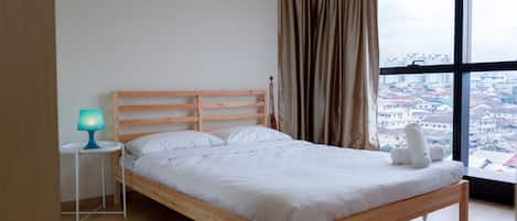 A bedroom with a wooden bed, a small bedside table with a lamp, a view of a cityscape through large windows, and a striped rug on the floor.