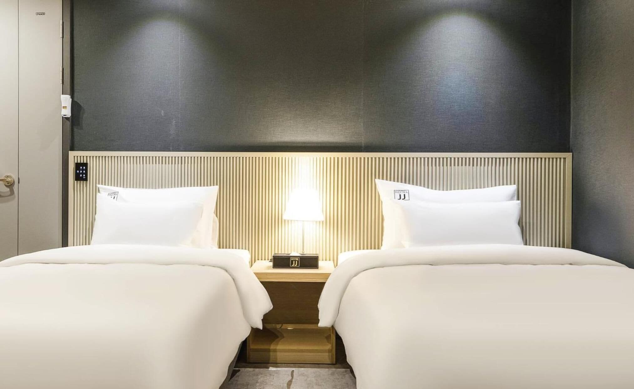 Una camera d'albergo con due letti, un comodino con una lampada e un pannello di controllo a parete.