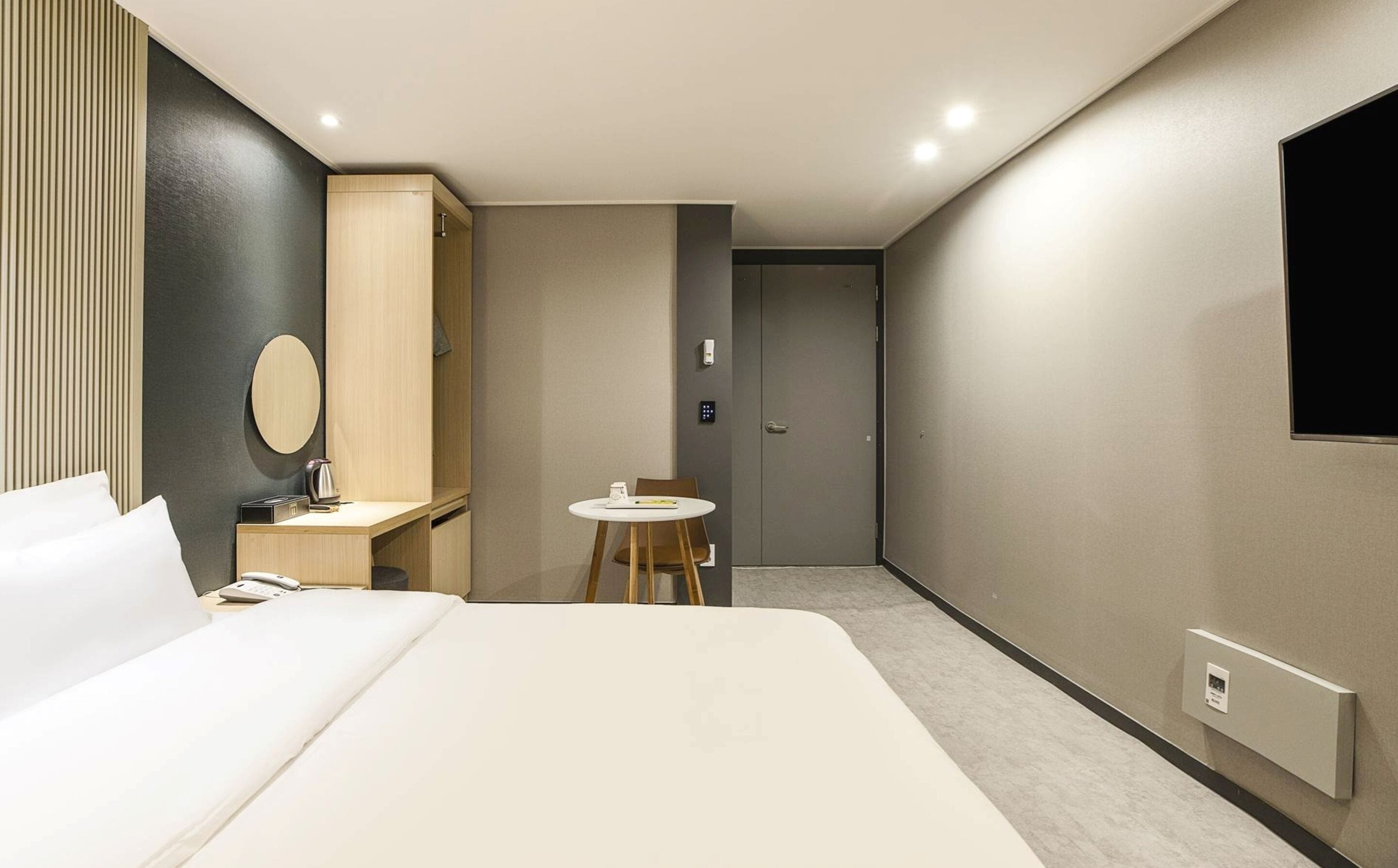 Une chambre d’hôtel moderne avec un grand lit, une table de chevet avec une lampe, une petite table ronde avec une chaise, une armoire, un miroir et un téléviseur fixé au mur.