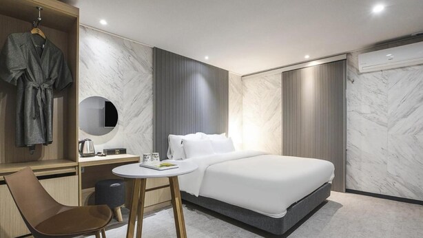 A modern hotel room with a bed, bedside table, chair, and a vanity with a mirror and robe. - JJ Hotel (Seongnam)