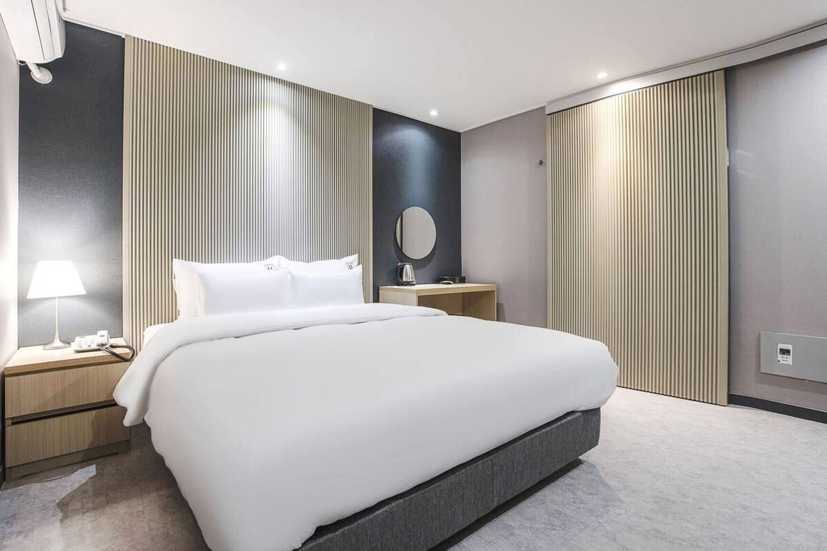 Une chambre d’hôtel moderne avec un grand lit, une table de chevet avec une lampe, un miroir rond et une petite table de chevet avec une bouilloire.
